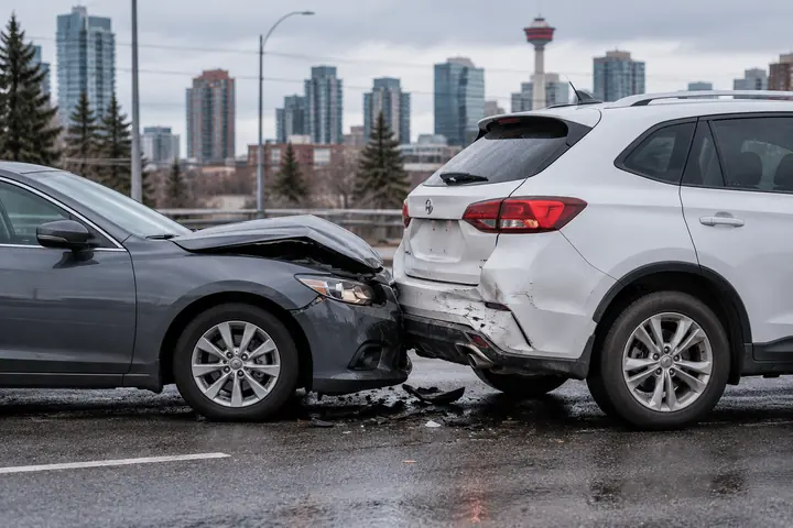 Grey sedan with a crumpled hood pressed into the rear bumper of a white SUV on a wet Calgary road, Calgary Tower in the background
